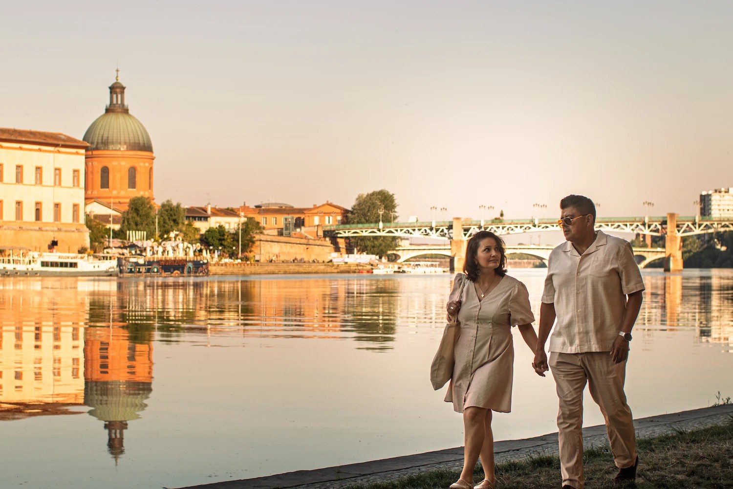 Couple holding hands along the Garonne in Toulouse at golden hour, with the dome of La Grave in the background